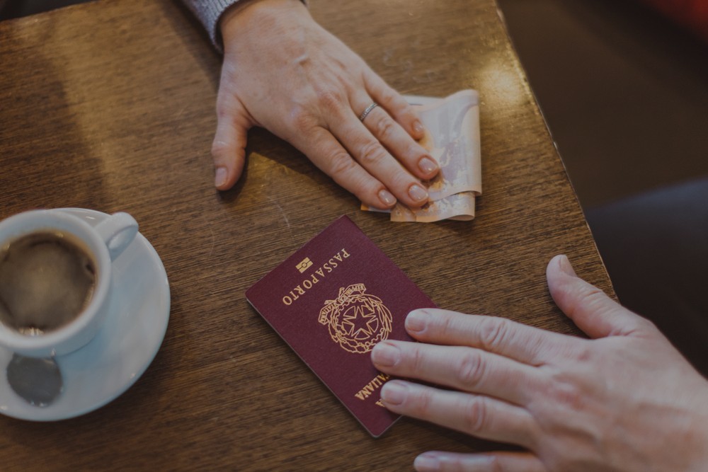 Image of two hands on a coffee table. There's a coffee next to a hand that sliding money towards another hand is sliding a passport back in exchange for the money.