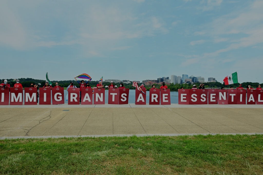Image of a pro-immigrant rally that features protestors holding red signs with white letters that read: "immigrants are essential."