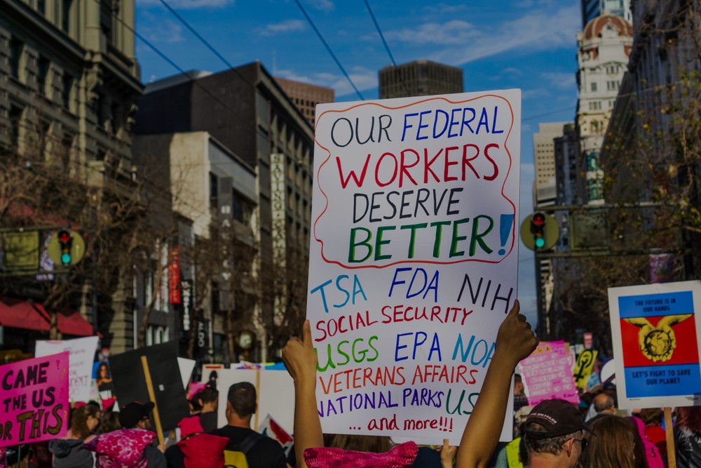 An image from a rally and protest against the massive layoffs of federal workers. One of them is holding a sign that reads "Our federal workers deserve better."
