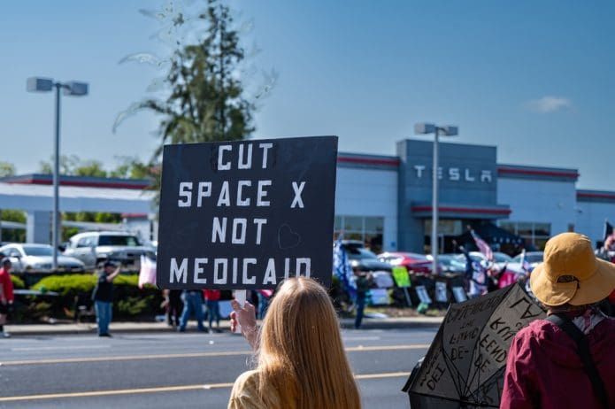 A demonstrator carries a sign reading "Cut Space X not Medicaid" outside a Tesla dealership.