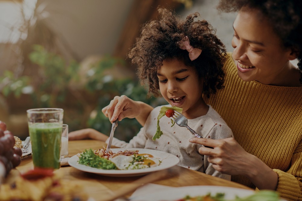 An image of a child seated on their mother's lap at a dinner table while being fed salad. Both of them are smiling
